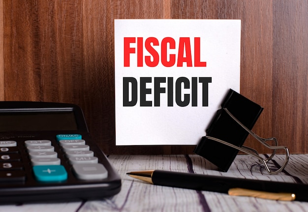 Déficit fiscal está escrito en una tarjeta blanca sobre un fondo de madera junto a una calculadora y un bolígrafo.