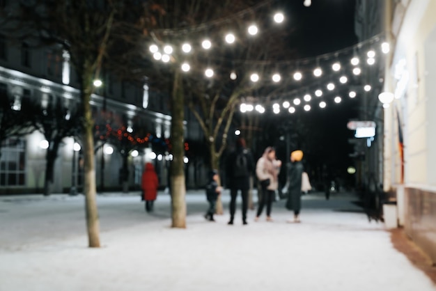 Defokussieren Sie die Stadtgasse mit Menschen, die am Winterabend im Freien von einer Girlande beleuchtet werden. Winter verschneite Straße, Park in Unschärfe, abstrakte Kulisse