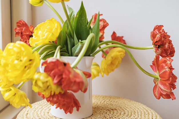 Defokussierte gelb-rot blühende Tulpenblumen mit grünen Stängeln und Blättern in weißem Keramikkrug auf hellweißem Wandhintergrund Kreative florale Botaniktapete Minimale kreative Grußkarte