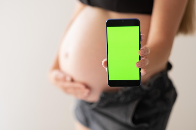 Defokussierte, nicht erkennbare schwangere Frau, die ihren Babybauch streichelt und ein Mobiltelefon mit grünem Bildschirm-Chromakey auf weißem Hintergrund hält