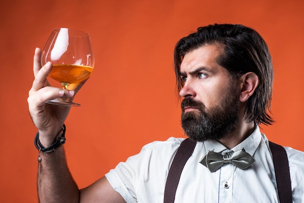 Degustación y bebida whisky escocés hombre barbudo en tirantes bebiendo ron vidrio elegante hombre de negocios usar pajarita para evento formal sommelier verdadero caballero con alcohol brandy o coñac