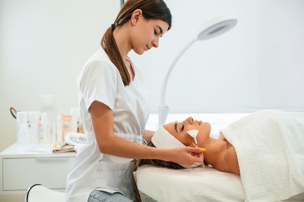 Deitada usando escovas rosto de mulher recebendo cuidado facial por mãos de esteticista em salão de spa