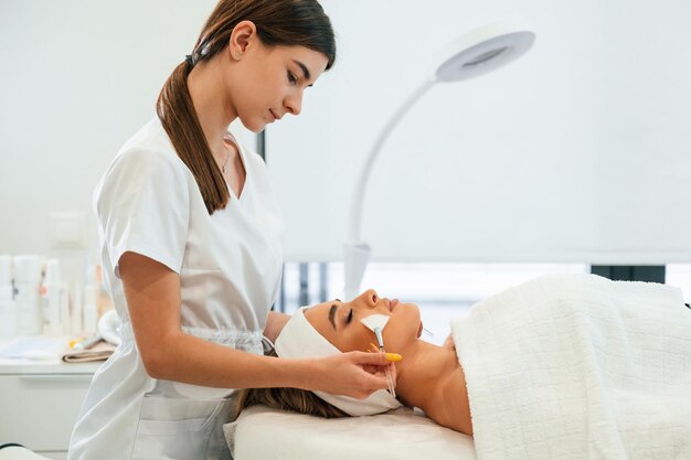 Foto deitada usando escovas rosto de mulher recebendo cuidado facial por mãos de esteticista em salão de spa