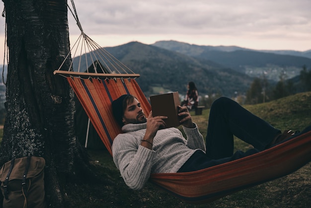 Deixe todas as suas preocupações para trás. Jovem bonito deitado na rede e lendo um livro enquanto acampa com a namorada