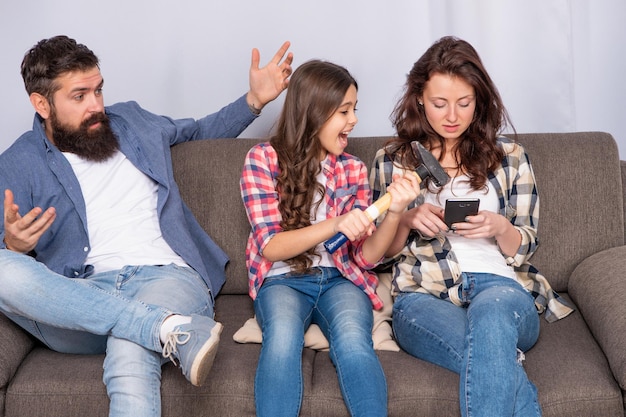 Deja de ignorarme. La madre usa el teléfono inteligente durante el tiempo en familia. Chica enojada amenaza con martillo