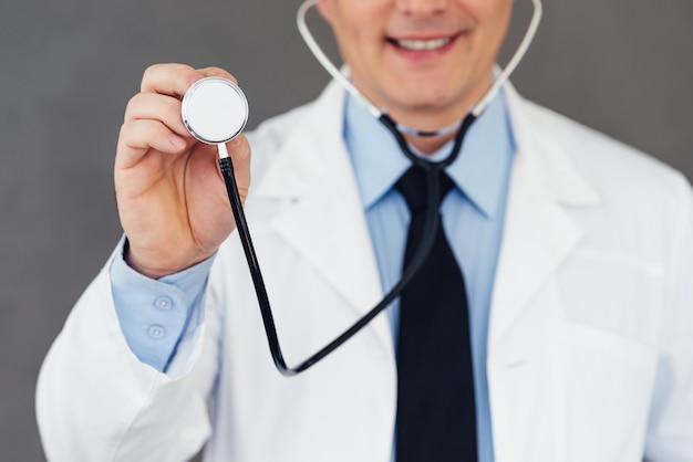 ¡Déjame escuchar los latidos de tu corazón! Parte cercana del médico varón maduro que sostiene el estetoscopio con una sonrisa mientras está de pie contra el fondo gris