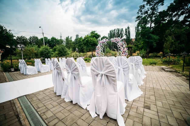 Dekoration einer Zeremonie im Freien in einem grünen Garten, ein Bogen aus frischen Blumen, ein weißer Weg für Jungvermählten, Stühle in weißen Stoffbezügen