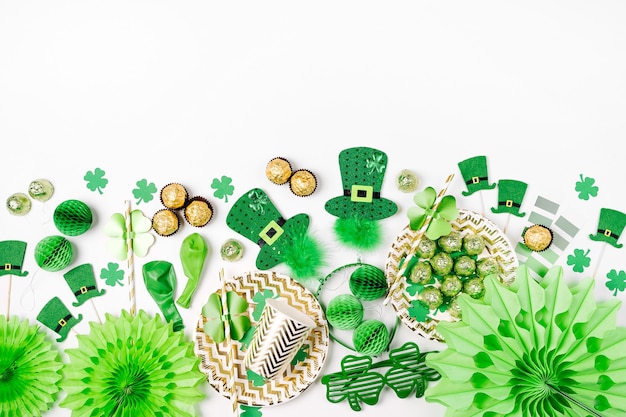 Dekorationen und Requisiten für die St. Patrick's Day Party. Grüne und goldene Papierdekorationen, Einwegteller und -becher, Hüte, Kleeblatt, Konfetti und Süßigkeiten auf weißem Hintergrund. Flache Lage, Ansicht von oben.