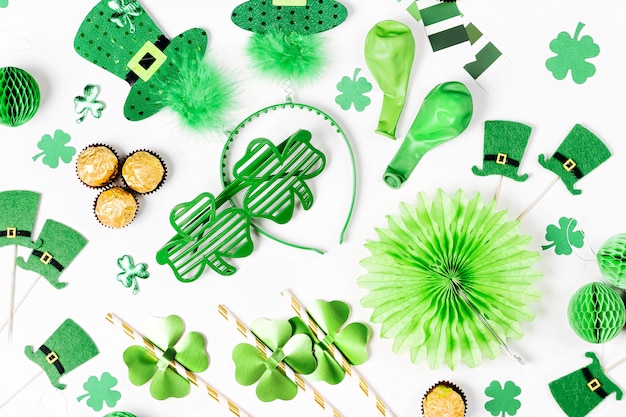 Dekorationen und Requisiten für die St. Patrick's Day Party. Grüne und goldene Papierdekorationen, Haarreifen mit Hüten, Kleeblatt, Sonnenbrillen, Luftballons, Konfetti und Süßigkeiten auf weißem Hintergrund. Flache Lage, Ansicht von oben