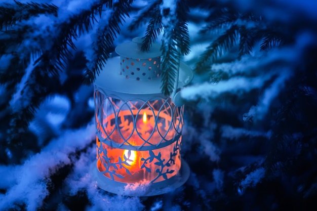 Dekorative Weihnachtslaterne mit brennender Kerze, die an einem schneebedeckten Tannenzweig in einem Winterpark hängt. Festliche Karte des neuen Jahres, Plakat, Postkartendesign.
