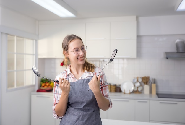 Con un delantal, una sonriente joven cocinera está de pie en la cocina, sosteniendo un cucharón y un batidor.