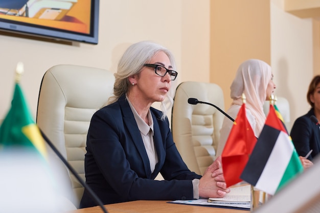 Delegada femenina madura seria en traje elegante escuchando preguntas de la audiencia antes de responderlas en la conferencia