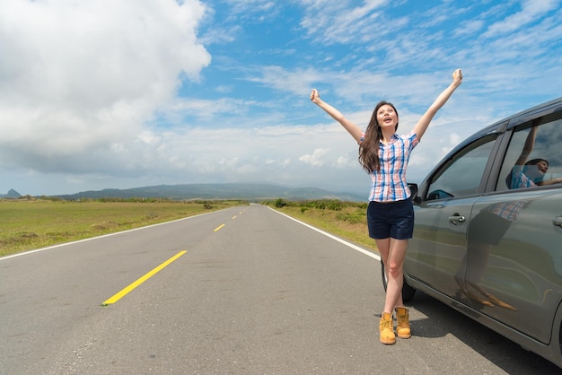 Foto delgada y elegante viajera que detiene el auto en la carretera de asfalto y se para al lado del auto levantando las manos disfrutando del paisaje rural local.