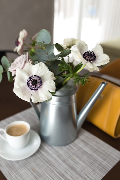 Delicadas flores de anémonas en un florero de metal con una taza de café sobre la mesa de madera, bolso de mano amarillo.