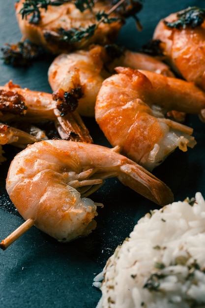 Deliciosa brocheta de camarones asados colocada sobre la superficie negra entre ajo asado y arroz blanco