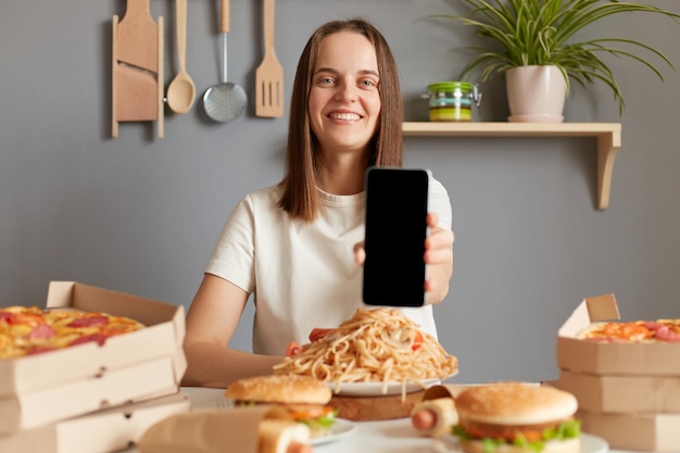 Deliciosa comida Pedidos en línea Entrega rápida Mujer sonriente con camiseta blanca sentada en la mesa con comida chatarra en la cocina que muestra el teléfono inteligente con espacio de copia de pantalla vacío para publicidad