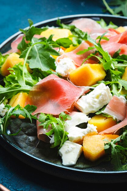 Deliciosa ensalada fresca de verano con melón melón prosciutto queso suave y rúcula en el espacio de copia de la vista superior del fondo de la mesa azul