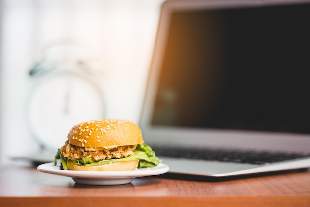 Una deliciosa hamburguesa en la mesa con una computadora portátil borrosa y un despertador