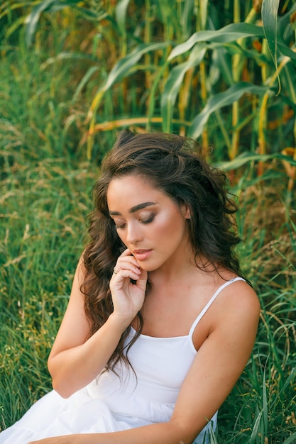 Deliciosa jovem em um vestido branco em um campo de milho verde na luz do sol Retrato de uma bela modelo com cabelo escuro longo e encaracolado Summer Harvest Eco