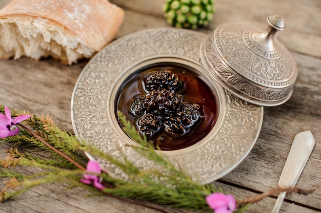 Foto deliciosa mermelada de conos en la placa de plata con una gorra, rama de árbol y pan cerca de él en la mesa de madera