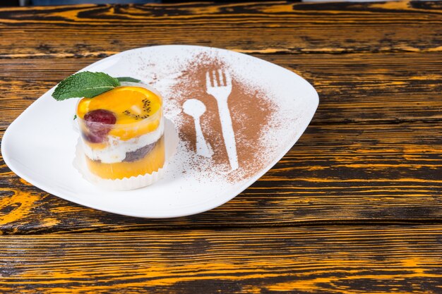 Deliciosa mousse de frutas cítricas com laranja e frutas vermelhas decorada com hortelã em um prato com silhuetas de um garfo e colher em cacau em pó, fundo de mesa de madeira rústica com copyspace