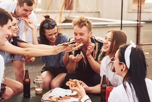 Deliciosa pizza grupo de jovens em roupas casuais fazem uma festa no telhado juntos durante o dia