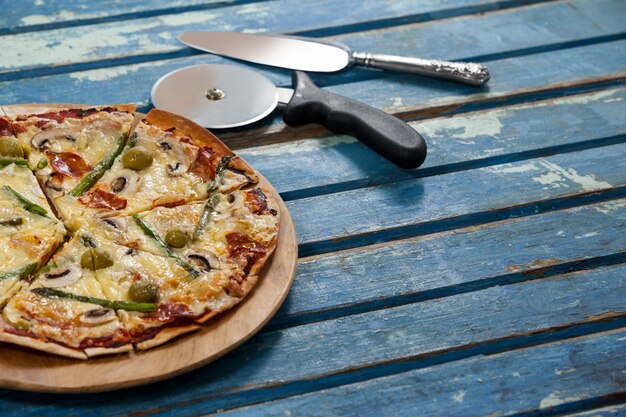 Deliciosa pizza servida en bandeja de pizza con cortador y cuchillo en tablón de madera