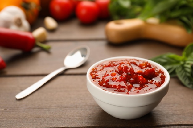 Deliciosa salsa adjika en tazón e ingredientes en primer plano de mesa de madera