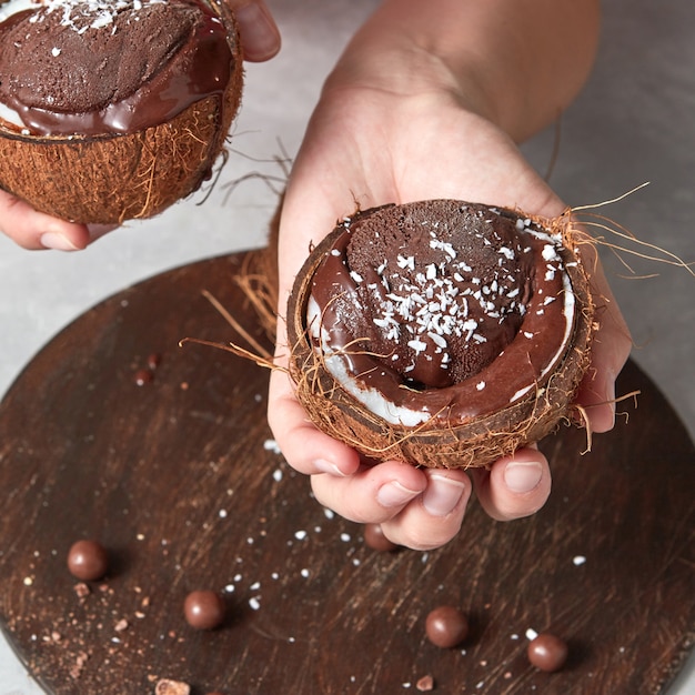Deliciosa sobremesa de chocolate derretido, sorvete em uma casca de coco nas mãos de uma garota acima de uma placa de madeira sobre uma mesa cinza.