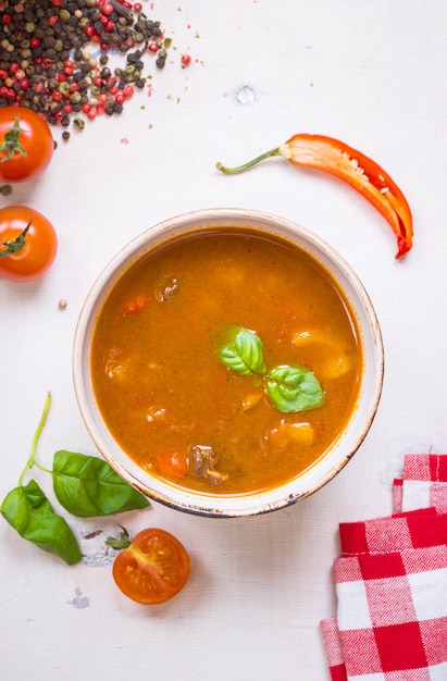 Deliciosa sopa de tomate com carne em uma tigela branca sobre uma mesa de madeira com tomate cereja fresco, folhas de manjericão, pimenta malagueta cortada e papel de cozinha guingão vermelho.