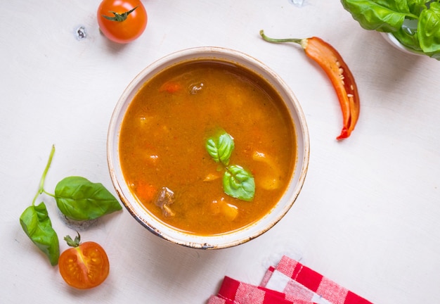 Deliciosa sopa de tomate com carne em uma tigela branca sobre uma mesa de madeira com tomate cereja fresco, folhas de manjericão, pimenta malagueta cortada e papel de cozinha guingão vermelho.