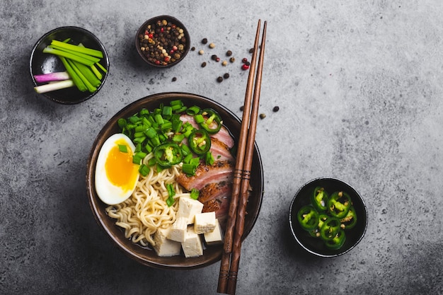 Deliciosa sopa de fideos asiáticos ramen en un tazón con caldo de carne, tofu, carne de cerdo en rodajas, huevo con yema, fondo de hormigón rústico gris, de cerca, vista superior. Sabrosa sopa de ramen japonesa caliente para cenar al estilo asiático