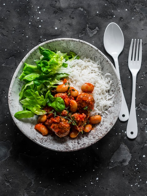 Deliciosas albóndigas equilibradas para el almuerzo con frijoles guisados en salsa de tomate con arroz y ensalada verde en una vista superior de fondo oscuro