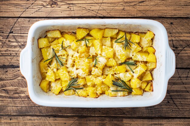 Deliciosas batatas assadas com especiarias, queijo e alecrim em uma assadeira de cerâmica sobre um fundo rústico de madeira.