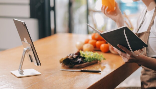 Deliciosas frutas y verduras en una mesa y mujer cocinando ama de casa está cortando pepinos verdes en una tabla de madera para hacer ensalada fresca en la cocina