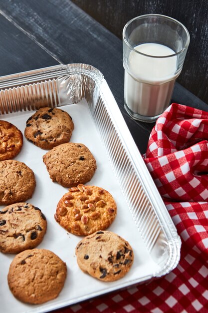 Deliciosas galletas naturales horneadas de avena - chocolate, nuez, con pasas en una bandeja para hornear con un vaso de leche y una toalla textil roja sobre una mesa de madera negra. Concepto de navidad.