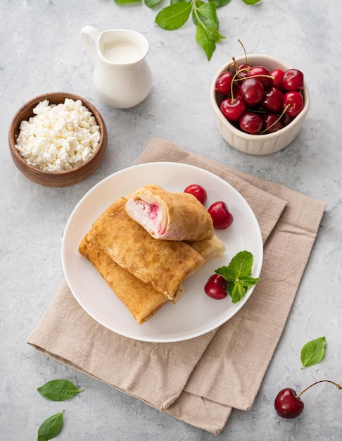 Deliciosas panquecas caseiras ou crepes com queijo cottage e cerejas frescas em um prato branco com creme azedo sobre fundo azul