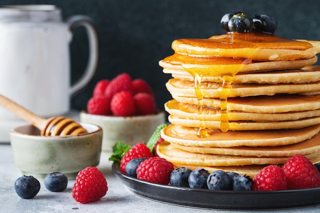 Deliciosas panquecas com mirtilos frescos, framboesa e xarope de ácer ou mel sobre fundo escuro