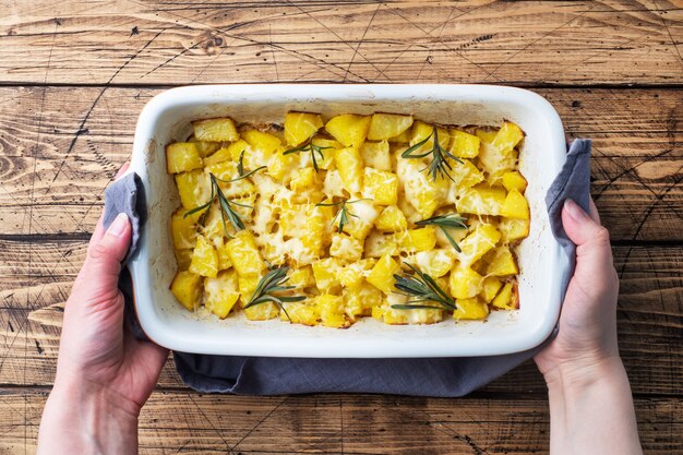 Deliciosas patatas al horno con especias, queso y romero en una bandeja para hornear de cerámica sobre un fondo rústico de madera.