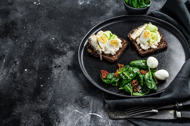 Deliciosas tapas de aperitivo, huevos pasados por agua y bocadillos de sardinas en lata. Ensalada de espinacas y tomates secos. Fondo negro.