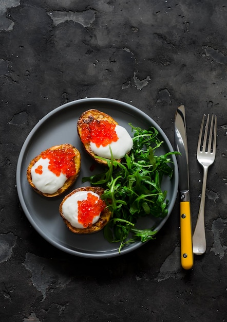 Foto deliciosas tapas aperitivo patatas a la parrilla con crema agria y caviar rojo y ensalada de rúcula en un fondo oscuro vista superior