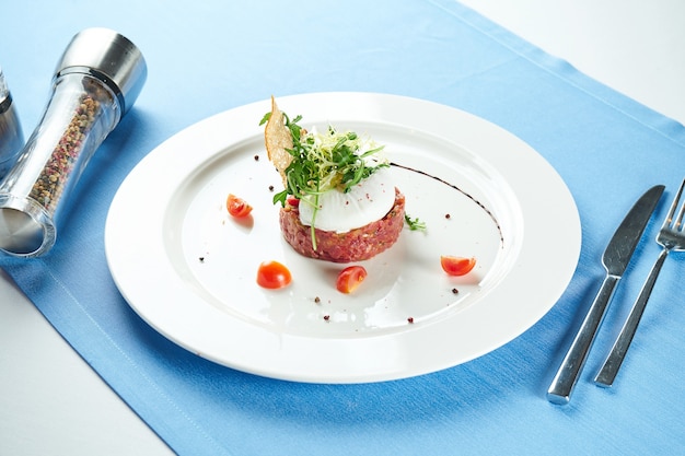 Delicioso aperitivo: tartar de ternera con picatostes, huevo escalfado y ensalada de rúcula en un plato blanco sobre un mantel azul