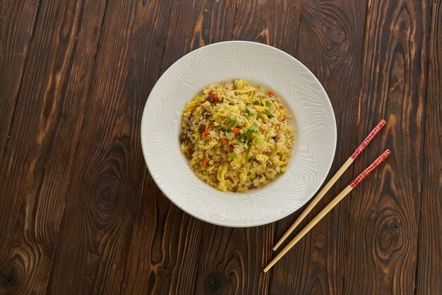 Delicioso arroz frito asiático com carne, ovo, cenoura, alho e cebola verde com pauzinhos vista horizontal de cima no prato branco de mesa de madeira, cópia espaço