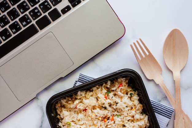 Delicioso arroz frito na lancheira na napery com colher de pau e garfo e computador portátil no local de trabalho