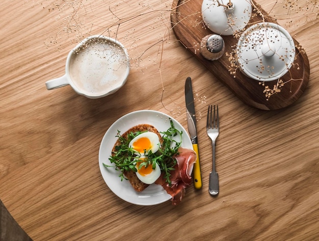 Delicioso brindis de desayuno con rúcula de huevo hervido y prosciutto y capuchino en una mesa de madera vista superior