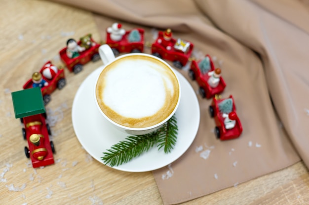 Delicioso café capuchino festivo fresco de la mañana en una taza de cerámica blanca sobre la mesa de madera con tren decorativo de navidad, adornos rojos, luciérnagas y ramas de abeto