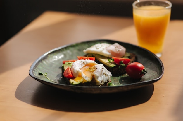 Delicioso café da manhã em casa. Sanduíche com abacate fresco fatiado acima de pão torrado de centeio com tomate cereja e ovo pochê na placa verde. A gema de suco de laranja fresco escorre na torrada