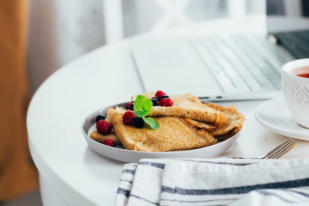 Delicioso café da manhã. mesa de trabalho branco com laptop e panquecas de crepes frescas, xícara de chá