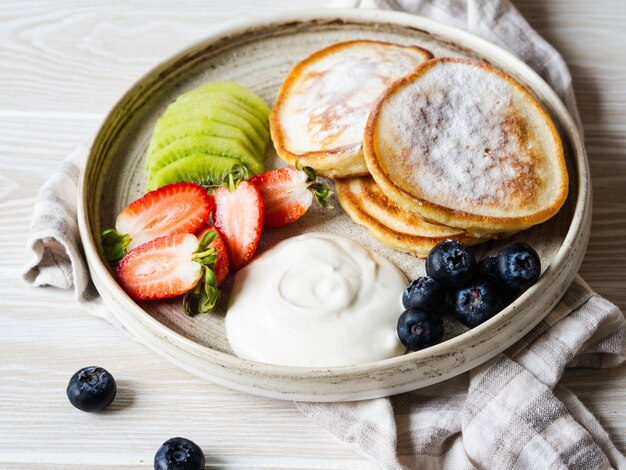 Delicioso desayuno: buñuelos de panqueques, espolvoreados con azúcar en polvo con frutas y bayas y crema agria en un plato sobre la mesa de madera. Copia espacio