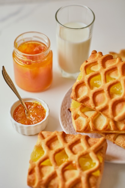 Delicioso desayuno con mini tartas de hojaldre con relleno de mermelada de pera y lima y un vaso de leche en la mesa blanca y tarro de mermelada